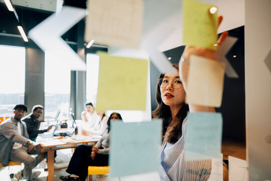 Young businesswoman explaining ideas on glass wall to coworkers sitting inside meeting room