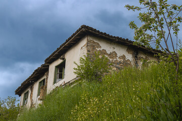 Fototapeta premium Old abandoned rural house on grassy hill under dramatic cloudy sky traditional architecture decay