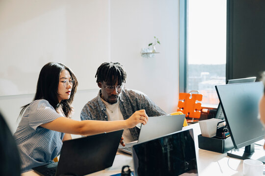 Young female programmer assisting male colleague working on laptop at creative workspace