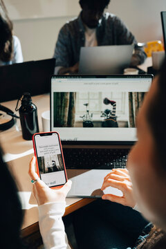 Over the shoulder view of businesswoman comparing websites on phone and laptop while working at office