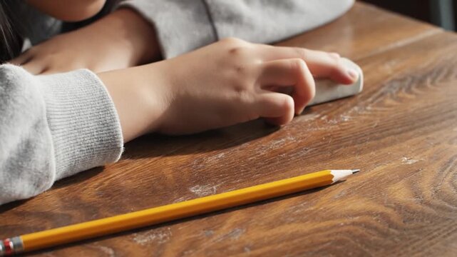 A diligent student's hand actively erases pencil marks from a textured wooden desk, illustrating the essential learning process of identifying and correcting errors during schoolwork or creative tasks