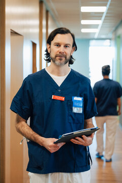 Portrait of confident male physician holding file folder while standing in hospital lobby