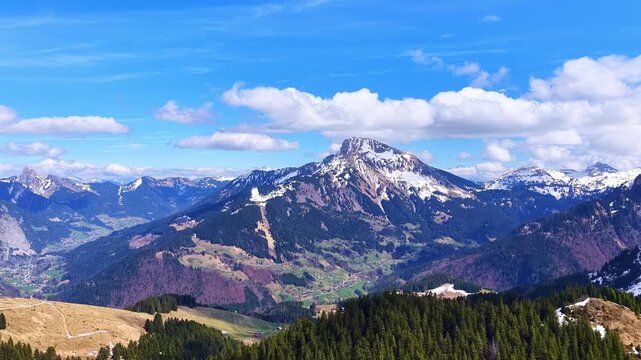 Drone 4K aerial forward flight over beautiful French Alps mountains with snowy peaks, forests and alpine valleys under blue sky in spring