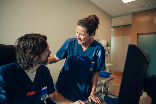 Smiling female medical expert talking with male coworker sitting at desk in office at hospital