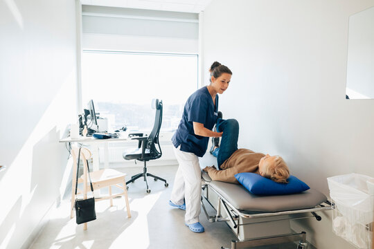 Female physiotherapist stretching leg of senior woman lying on examination table in hospital