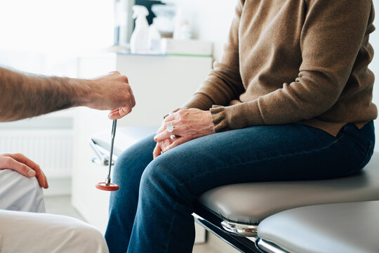 Midsection of male doctor checking patient's knee with reflex hammer in examination room at hospital