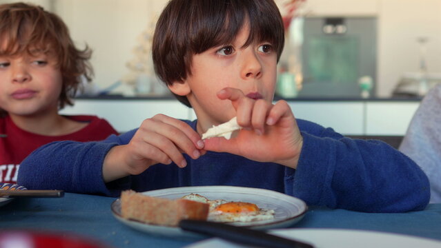 Boy eating fried egg with bread at breakfast table, using hands to take bite of food while sitting with siblings during family meal at home