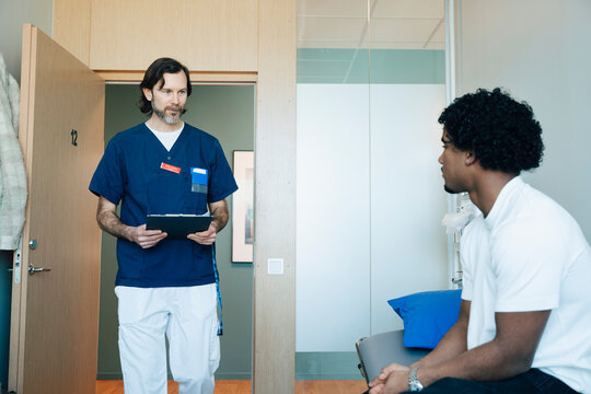 Male doctor wearing scrubs walking towards young patient sitting on table in examination room at clinic