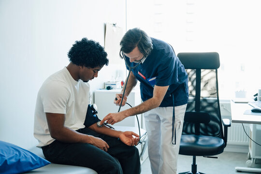 Male physician checking blood pressure of young man while using stethoscope on arm during medical exam in room at hospit
