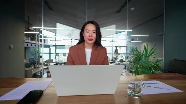 Young professional specialist Asian business lady working on laptop pc sitting at desk in modern office space. Middle-aged eastern mature woman using computer technology app for work online. Push in