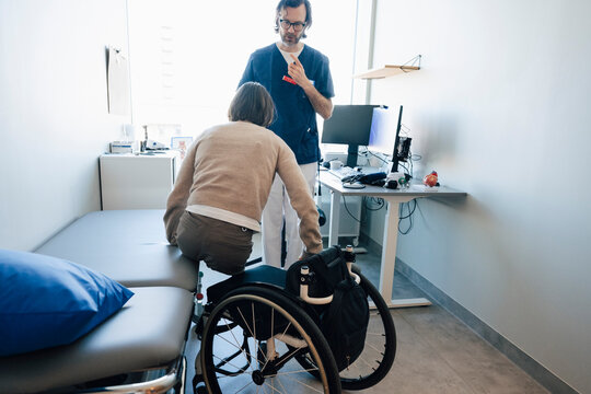 Male doctor talking with female amputee woman sitting on wheelchair from table in examination room at hospital