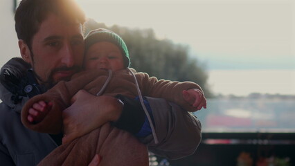 Father making playful face while holding baby in bear teddy fleece coat and striped beanie hat outdoors, man bonding with child during joyful cold season family outing © Marco