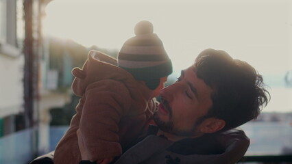 Father making playful face while holding baby in bear teddy fleece coat and striped beanie hat outdoors, man bonding with child during joyful cold season family outing © Marco