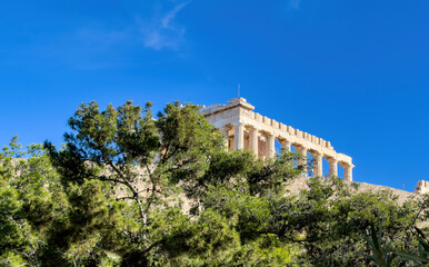 Naklejka premium Parthenon temple on the Acropolis hill in Athens, Greece. Stone walls and green trees, blue sky, sunny day