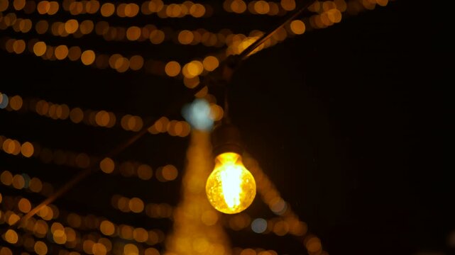 Golden christmas lights string glowing at night. Close up view of a glowing yellow light bulb swaying on a string. Blurry christmas lights and a decorated tree create a beautiful golden bokeh effect