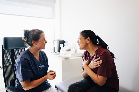 Smiling woman showing gratitude towards female doctor while sitting on table in clinic