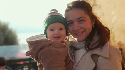 Happy baby smiling in bear teddy fleece coat and striped beanie hat while mother embraces outdoors, family enjoying cold season together, parent and child sharing warmth and love © Marco