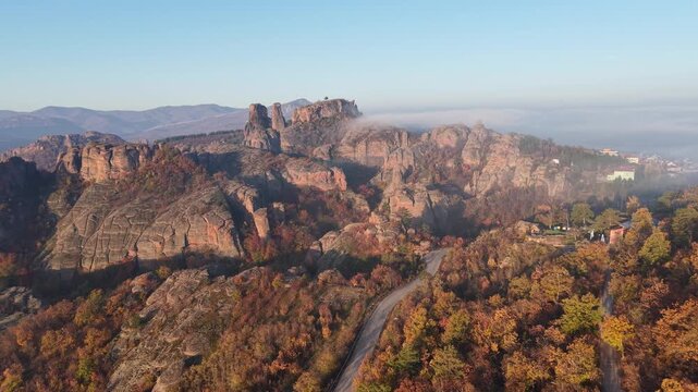 Wide aerial view of a large natural rock massif above autumn forest with low fog drifting across the upper ridge.