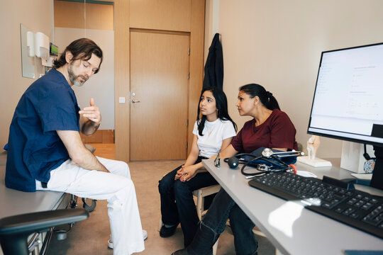Male pediatrician explaining condition to mother and daughter while sitting on examination table in clinic
