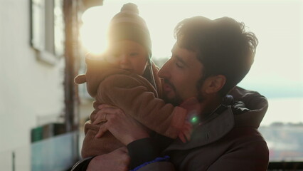 Father holding baby in bear teddy fleece coat and striped green beanie hat outdoors, man smiling with joy as infant hugs his face during warm and affectionate winter moment © Marco