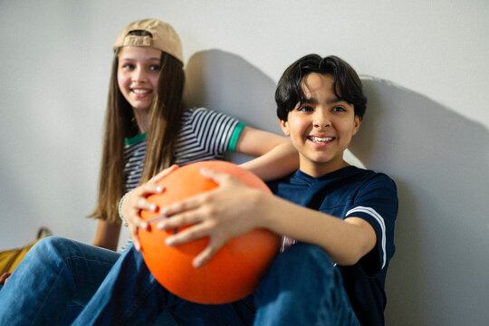 Happy siblings in casual clothes looking away while sitting with basketball by wall