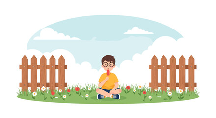 Fototapeta premium Boy with glasses sitting on the grass and enjoying a red popsicle in a beautiful field with flowers and a wooden fence.