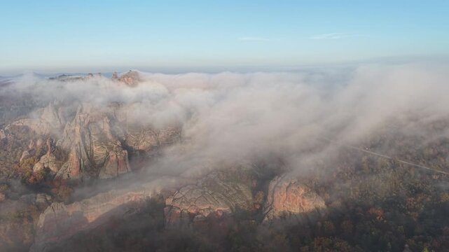Wide aerial view of natural rock formations partially covered by dense low fog above colorful autumn woodland.