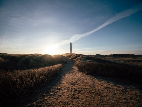 Sandy path winding through coastal dunes toward a lighthouse at sunrise in Denmark, with warm golden light illuminating the rugged seaside landscape.