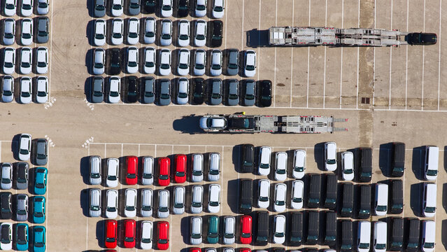 New cars, vans, and utility vehicles are parked in an organized outdoor storage lot, awaiting transportation by semi-trailer car carriers for distribution to dealerships