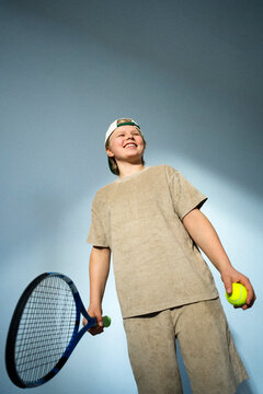 Low angle view of smiling boy holding tennis racket and ball against blue background