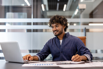 Fototapeta premium Smiling Indian man in the office at the table working with documents and typing on a laptop