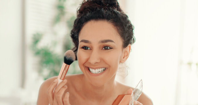 A woman is applying makeup in a bright room. She smiles while holding a brush in one hand and a compact in the other. The setting has soft lighting with greenery in the background.