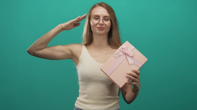 Young blonde woman wearing glasses salutes with raised bare arm while holding pink ribboned gift box in studio; playful celebration.