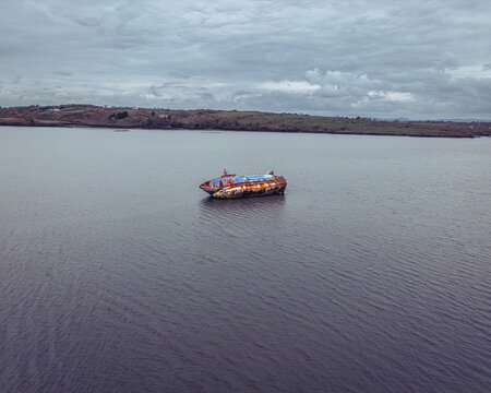 Aerial view of a rusty abandoned shipwreck floating in the calm waters of a wide river with green hills along the shoreline under an overcast sky.