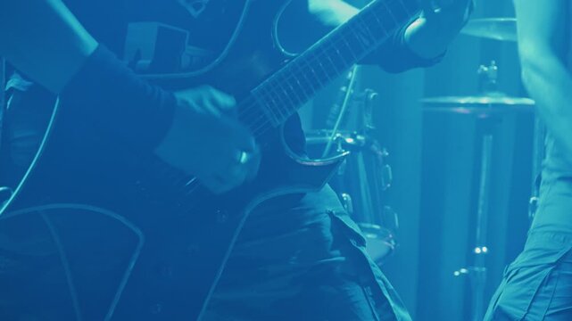 Close up of guitarist's hands playing fast riff on electric guitar. Stage lit with green lights creates intense atmosphere for heavy metal concert. Band members and drums visible in background