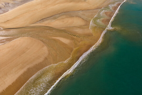 Aerial view of the sandy beach with intricate water patterns and turquoise sea waves breaking on the shore in Plurien, Brittany, France.