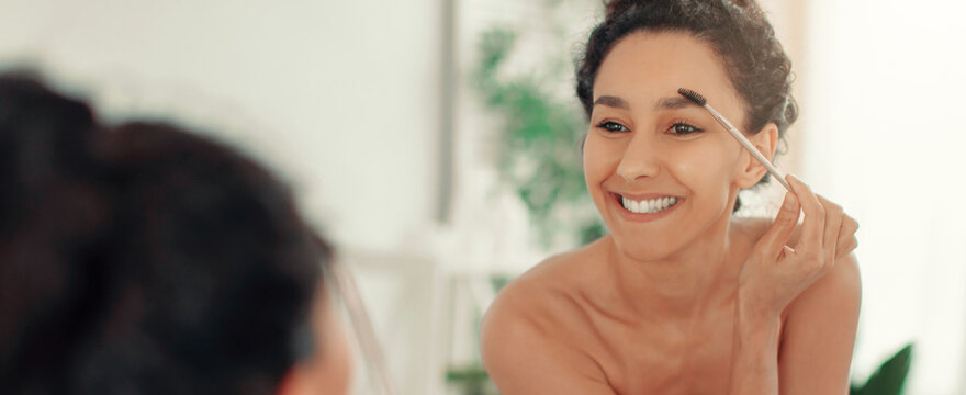 A woman smiles as she uses a brush to groom her eyebrows while looking into a mirror. The setting is a brightly lit bathroom with a relaxed atmosphere.