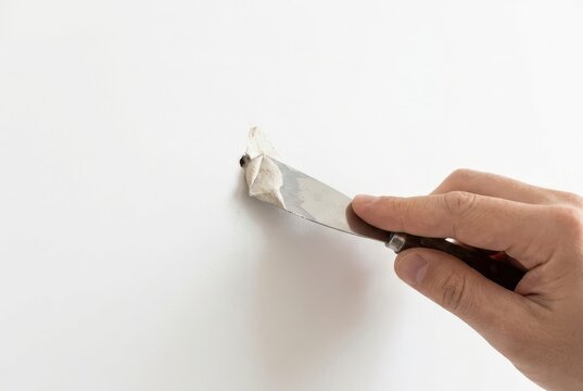 Close-up of a hand using a putty knife to repair a small hole in a white wall surface.