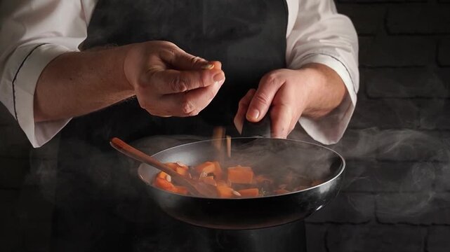 Chef in black apron adds seasoning to sauteed vegetables in a frying pan, steam rising against a textured brick wall backdrop