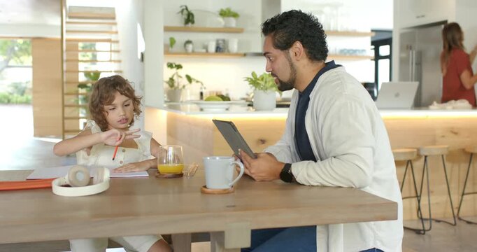 Child at kitchen table pausing with red pencil writing work while father watching tablet