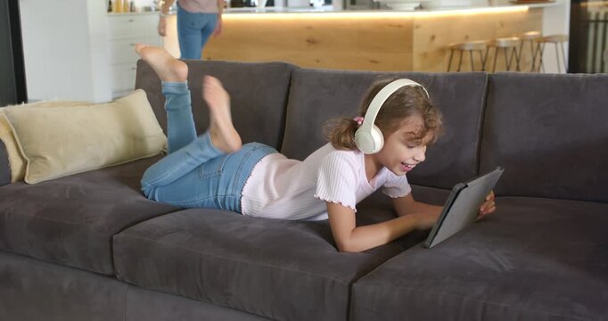 Female child lying on sofa in white headphones, tablet drawing focus tapping as parents making food