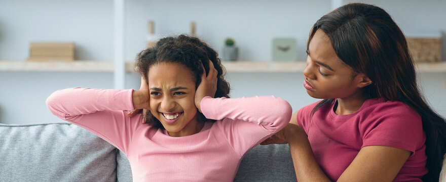 A mother talks to her daughter who covers her ears and looks upset. They sit on a couch in a living room with simple decor. The mother tries to help her daughter feel better during a hard talk.