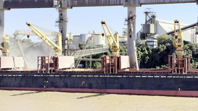 cargo ship with cranes moored to grain terminal. loading wheat by shore pipes in progress. view from sea. 