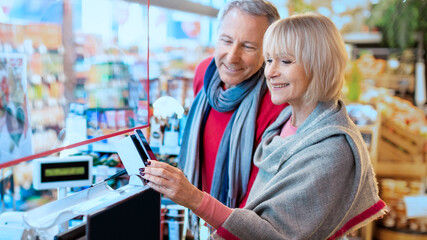 A couple stands close together in a grocery store. They are reviewing a mobile device. The store shelves are stocked with various items. Bright lights illuminate the scene. © Prostock-studio