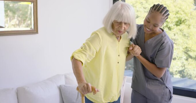 Carer in gray scrubs reaching wrist steadying as senior shifting, standing with cane in living room