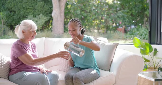 Caregiver woman and senior woman smiling as caregiver pouring from clear pitcher into glass on sofa
