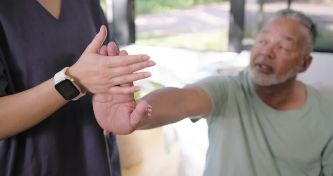 Asian caregiver in scrubs with smartwatch supporting senior man reaching in sunroom guiding fingers
