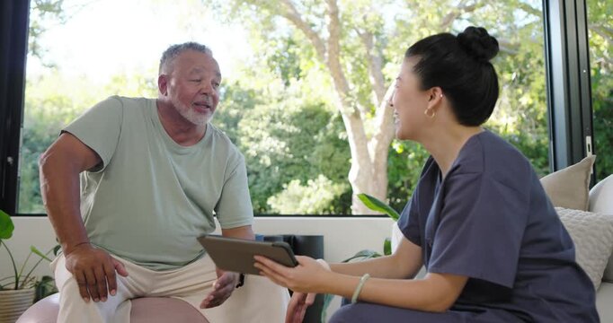 Asian nurse in scrubs showing tablet and explaining care at home while man smiling, fist-bump