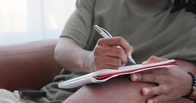 African American man writing on notebook with pen on couch in green shirt, pausing, checking phone