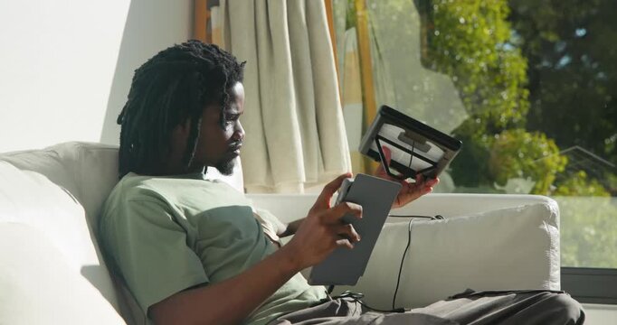 African American man on sofa reacting to sun, angling solar panel to window to charge tablet